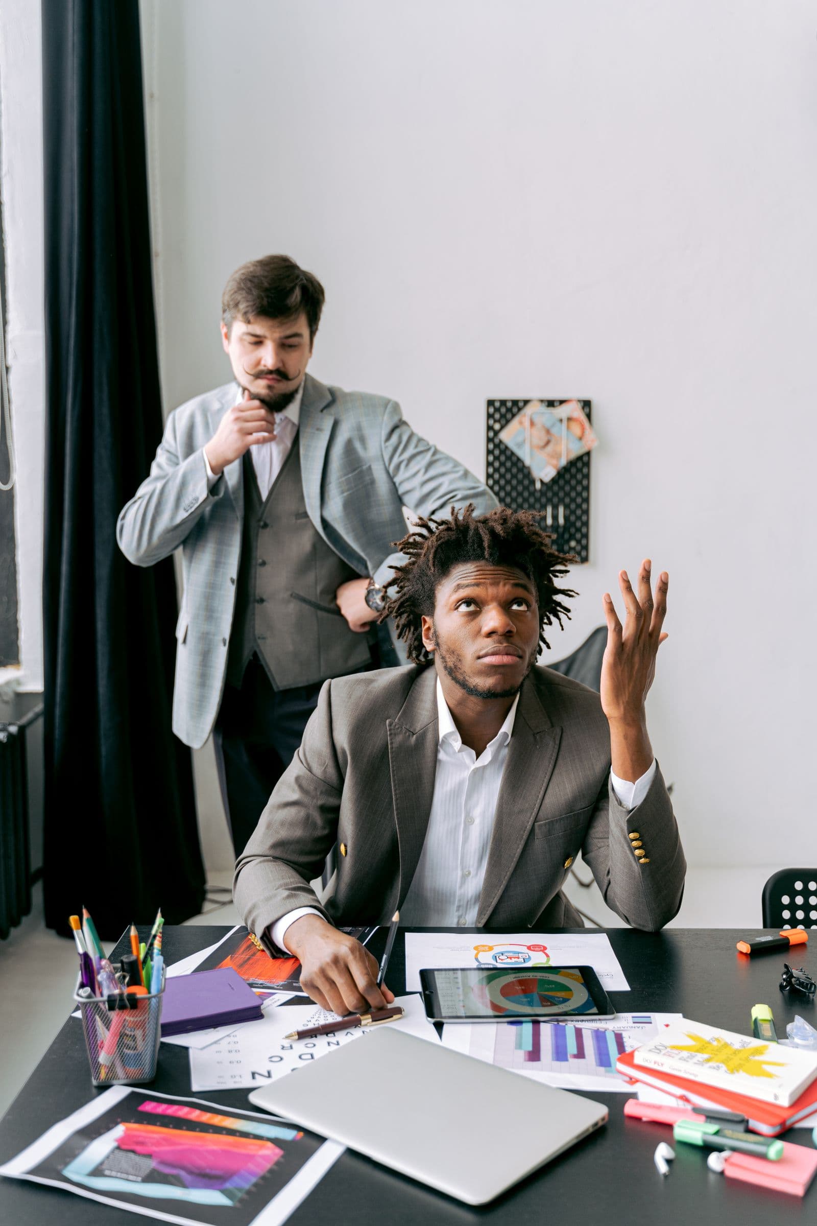 Two businessmen facing work challenges at a desk filled with papers and technology, conveying stress and contemplation.