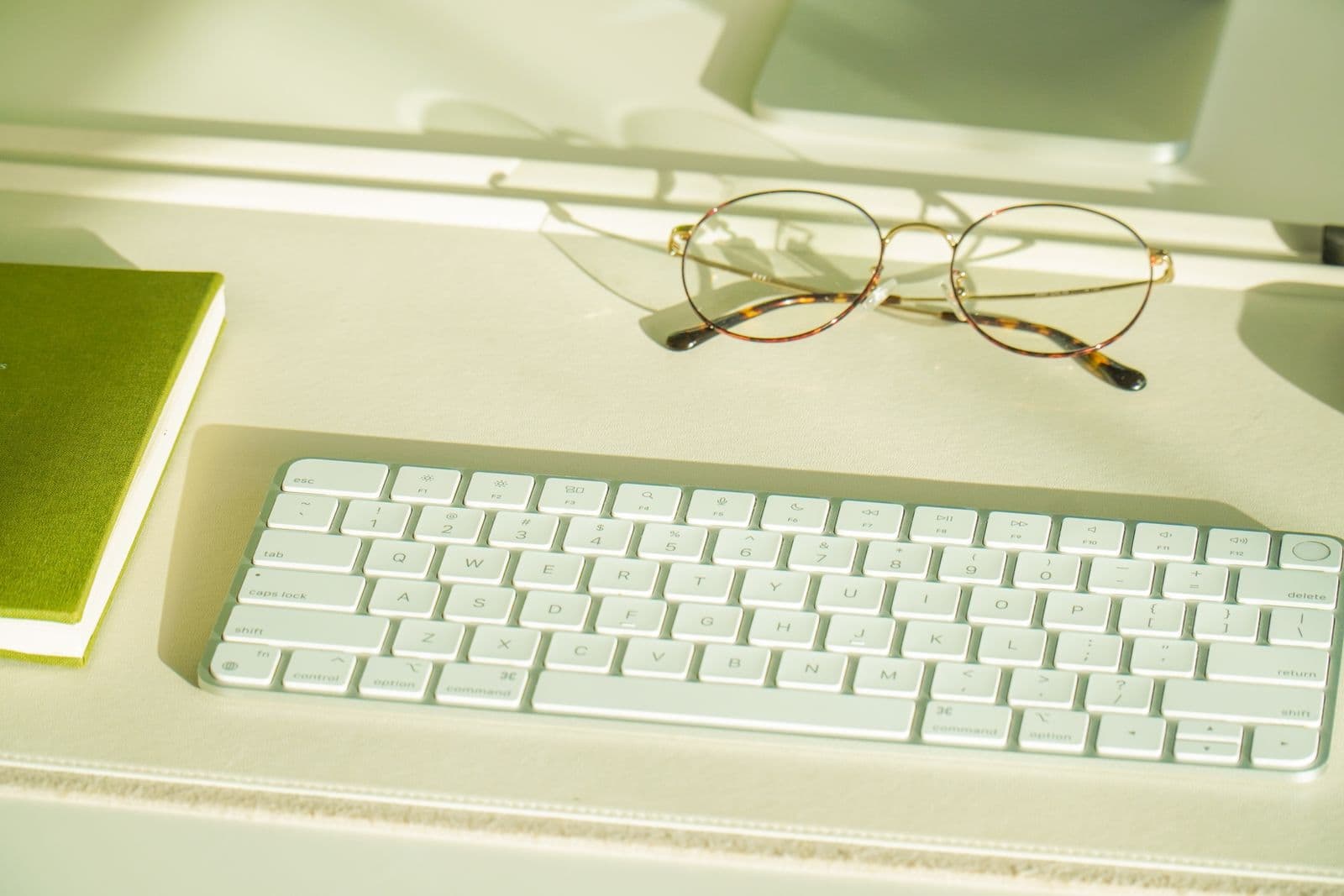 Sleek desk setup featuring keyboard, green notebook, and glasses. Ideal for productivity themes.